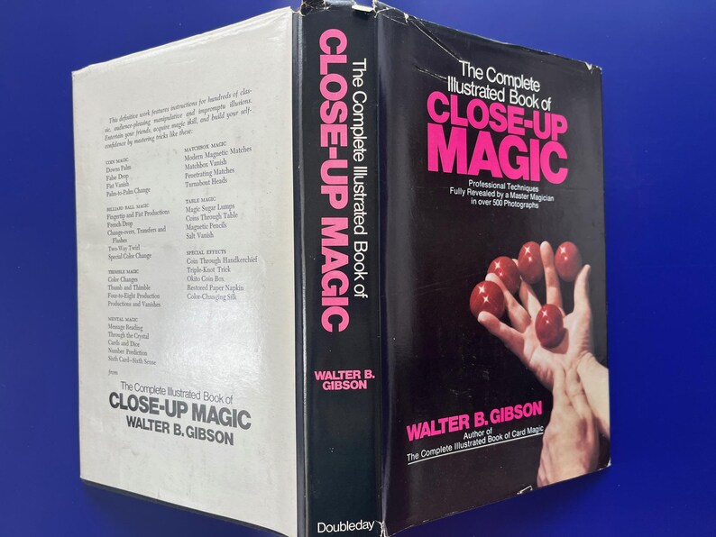 May include: A vintage book titled "The Complete Illustrated Book of Close-Up Magic" by Walter B. Gibson. The cover features a hand holding red balls. The spine displays the title in pink letters. The book is on a blue surface.