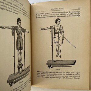 K&ouml;nnte beinhalten: Ein offenes Buch mit Illustrationen aus "Modern Magic". Die Zeichnungen zeigen Figuren, die scheinbar schweben oder Illusionen vollf&uuml;hren. Der Text enth&auml;lt Ma&szlig;e in Fu&szlig; und Zoll sowie Beschreibungen der Tricks.