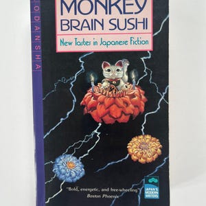 May include: A book titled "Monkey Brain Sushi: New Tastes in Japanese Fiction" with a black cover and a pink banner. The cover features a cat figurine, flowers, and lightning bolt designs. The book is from Kodansha.