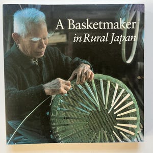 May include: Book cover showing an elderly man crafting a basket. The title reads "A Basketmaker in Rural Japan." The man is wearing a dark jacket and is focused on his work. The basket is green and white.