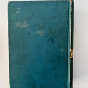 K&ouml;nnte beinhalten: Ein Vintage-Buch mit t&uuml;rkisblauem Einband und dunklem R&uuml;cken. Der Einband weist Gebrauchsspuren auf, mit verblassten Flecken und einer abgenutzten Ecke. Der R&uuml;cken hat ein beiges Etikett. Das Buch steht vor einem wei&szlig;en Hintergrund.