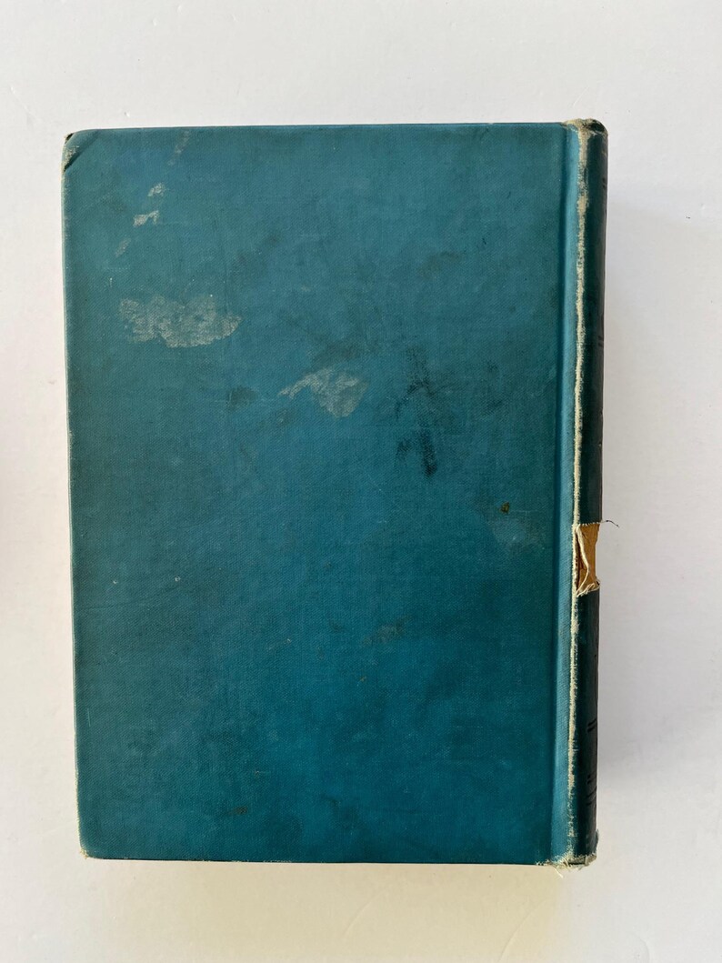 K&ouml;nnte beinhalten: Ein Vintage-Buch mit t&uuml;rkisblauem Einband und dunklem R&uuml;cken. Der Einband weist Gebrauchsspuren auf, mit verblassten Flecken und einer abgenutzten Ecke. Der R&uuml;cken hat ein beiges Etikett. Das Buch steht vor einem wei&szlig;en Hintergrund.