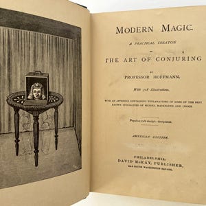 K&ouml;nnte beinhalten: Ein altes Buch mit dem Titel "Modern Magic" und einer Illustration eines Zaubertricks. Die Abbildung zeigt eine Kiste auf einem Tisch mit dem Kopf einer Person darin. Es ist eine amerikanische Ausgabe.