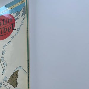 May include: A vintage book cover for "The Adventures of Tintin in Tibet." The cover features a red circle with white text, a snowy mountain scene, and footprints. The book's spine is visible, showing green and white details.