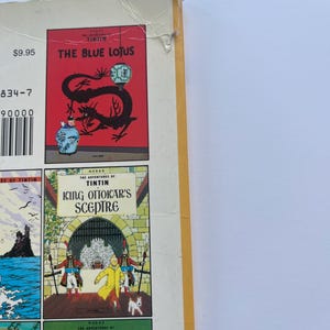 May include: A collection of Tintin comic books. The cover of "The Blue Lotus" features a red background with a black dragon and a blue vase. Other titles include "King Ottokar's Sceptre" and "The Seven Crystal Balls". The books have a white spine and yellow edges.