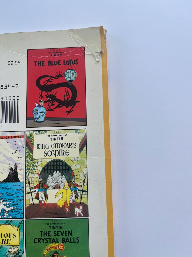 May include: A collection of Tintin comic books. The cover of "The Blue Lotus" features a red background with a black dragon and a blue vase. Other titles include "King Ottokar's Sceptre" and "The Seven Crystal Balls". The books have a white spine and yellow edges.