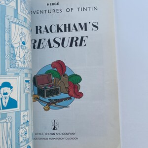 May include: A vintage copy of "The Adventures of Tintin: Red Rackham's Treasure" by Herg&eacute;. The cover features a colorful illustration of a treasure chest, feathers, and a pistol. The book is published by Little, Brown and Company.