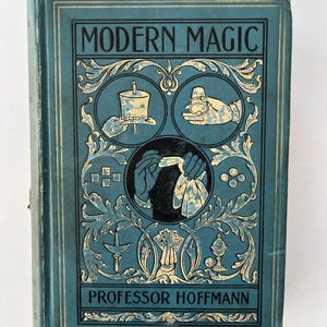 K&ouml;nnte beinhalten: Antikes Buch mit dem Titel "Modern Magic" von Professor Hoffmann. Der Einband ist t&uuml;rkis mit goldenen Akzenten und Illustrationen von Zaubertricks. Das Buch hat ein abgenutztes Aussehen.