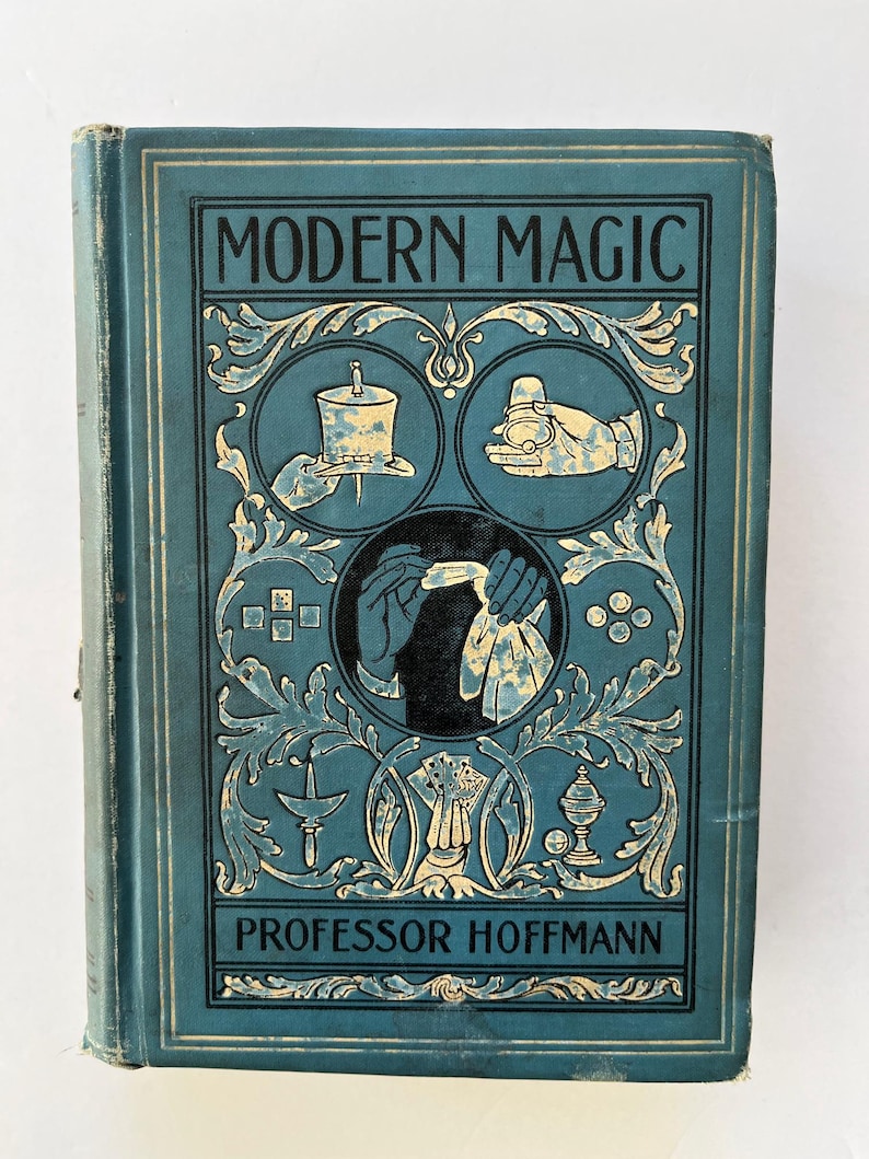 K&ouml;nnte beinhalten: Antikes Buch mit dem Titel "Modern Magic" von Professor Hoffmann. Der Einband ist t&uuml;rkis mit goldenen Akzenten und Illustrationen von Zaubertricks. Das Buch hat ein abgenutztes Aussehen.