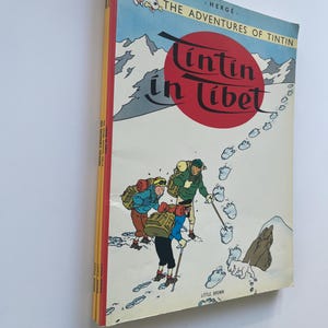 May include: A stack of vintage comic books featuring the cover of "Tintin in Tibet" by Herg&eacute;. The cover art depicts a snowy mountain scene with cartoon characters and the title in red. The books have a red spine.