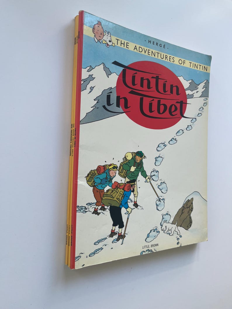 May include: A stack of vintage comic books featuring the cover of "Tintin in Tibet" by Herg&eacute;. The cover art depicts a snowy mountain scene with cartoon characters and the title in red. The books have a red spine.
