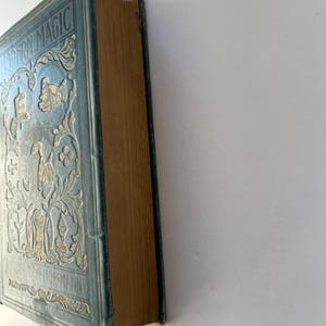 K&ouml;nnte beinhalten: Ein Vintage-Buch mit dem Titel "MODERN MAGIC" mit hellblauem Einband und goldgepr&auml;gten Designs. Der Buchr&uuml;cken ist sichtbar und zeigt gealterte, hellbraune Seiten. Das Cover zeigt verzierte Blumen- und Figurenillustrationen.