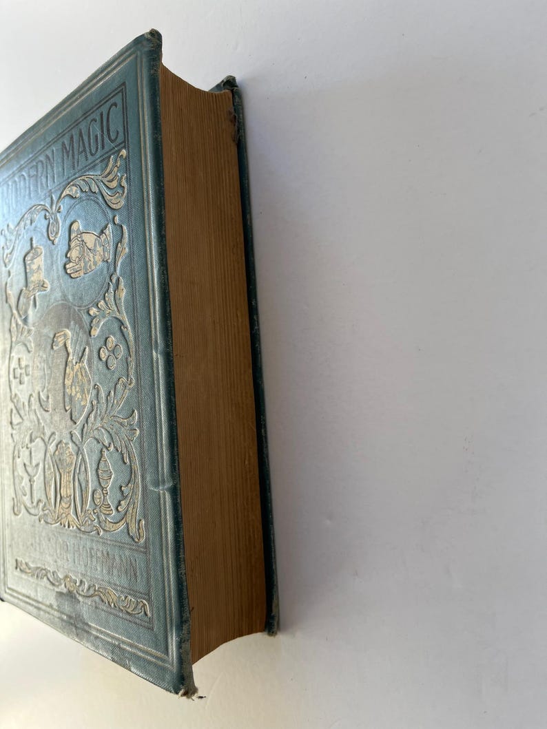 K&ouml;nnte beinhalten: Ein Vintage-Buch mit dem Titel "MODERN MAGIC" mit hellblauem Einband und goldgepr&auml;gten Designs. Der Buchr&uuml;cken ist sichtbar und zeigt gealterte, hellbraune Seiten. Das Cover zeigt verzierte Blumen- und Figurenillustrationen.