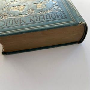 K&ouml;nnte beinhalten: Ein antikes Buch mit dem Titel "MODERN MAGIC" mit einem t&uuml;rkisblauen Einband und goldgepr&auml;gten Details. Die Seiten des Buches sind gealtert, und der Einband weist Abnutzungserscheinungen auf. Das Buch befindet sich vor einem wei&szlig;en Hintergrund.