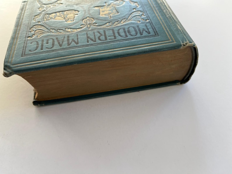 K&ouml;nnte beinhalten: Ein antikes Buch mit dem Titel "MODERN MAGIC" mit einem t&uuml;rkisblauen Einband und goldgepr&auml;gten Details. Die Seiten des Buches sind gealtert, und der Einband weist Abnutzungserscheinungen auf. Das Buch befindet sich vor einem wei&szlig;en Hintergrund.