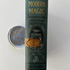 K&ouml;nnte beinhalten: Ein Vintage-Buch in T&uuml;rkis mit dem Titel "Modern Magic" von Professor Hoffmann, mit goldfarbenen Buchstaben und Illustrationen. Das Buch weist Gebrauchsspuren auf, mit einem zerrissenen Abschnitt auf dem Einband. Ein silberfarbenes, rundes Objekt liegt neben dem Buch.