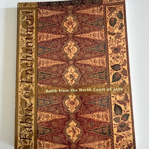 以下が含まれることがあります： 「Fabric of Enchantment」というタイトルのハードカバー本で、「Batik from the North Coast of Java」というサブタイトルが付いています。表紙には、赤、金、ティール色の詳細なバティック模様が施されており、花柄と幾何学模様が特徴です。
