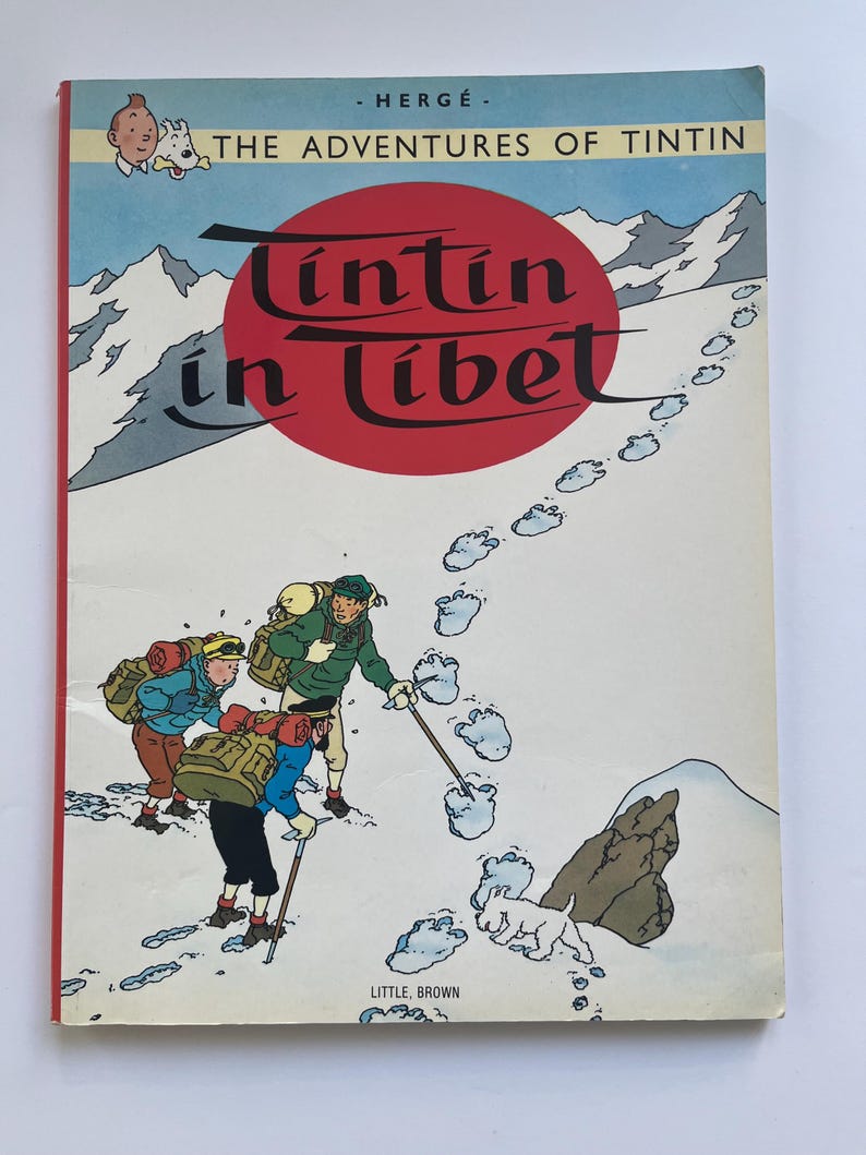 May include: A vintage copy of "The Adventures of Tintin in Tibet" comic book. The cover features a red circle with the title in white script, and illustrations of snowy mountains, footprints, and cartoon characters. Published by Little, Brown.