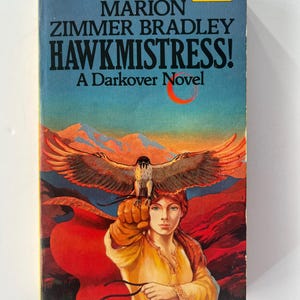 May include: A vintage paperback book titled "HAWKMISTRESS!" by Marion Zimmer Bradley. The cover art depicts a woman with a falcon on her gloved hand, set against red mountains and a blue sky. The book is a "Darkover Novel."
