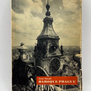 May include: A vintage book cover depicting a detailed illustration of a clock tower with a spire. The book, titled "Baroque Prague" by Arne Novák, shows signs of wear, indicating its age and history.