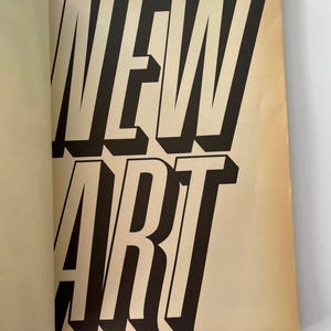 K&ouml;nnte beinhalten: Offene Buchseite mit den Worten "NEW ART" in gro&szlig;en, fetten, schwarzen Buchstaben mit wei&szlig;er Umrandung. Die Buchstaben haben einen 3D-Effekt und der Hintergrund ist hellbraun. Die linke Seite des Buches ist sichtbar.