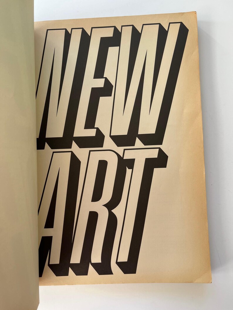 K&ouml;nnte beinhalten: Offene Buchseite mit den Worten "NEW ART" in gro&szlig;en, fetten, schwarzen Buchstaben mit wei&szlig;er Umrandung. Die Buchstaben haben einen 3D-Effekt und der Hintergrund ist hellbraun. Die linke Seite des Buches ist sichtbar.