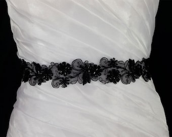 May include: A white dress with a black beaded and lace sash. The sash features a floral design with black beads.