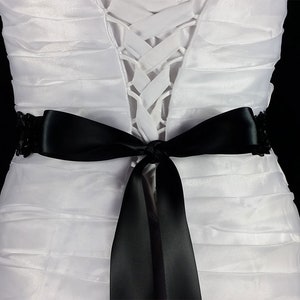 May include: A white dress with a black satin ribbon tied in a bow at the waist. The ribbon has a beaded embellishment on each end.
