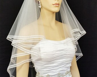 May include: A white bridal veil with a satin edge. The veil is layered and has a single tier. The veil is worn by a mannequin with red hair.