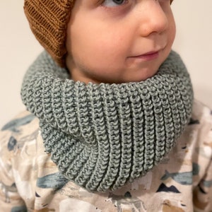 Könnte beinhalten: Ein Kind trägt eine braune Strickmütze mit einem weißen Blattlogo und einen salbeigrünen Strick-Halswärmer. Das Kind trägt ein gemustertes Hemd mit weißem Hintergrund und braunen, blauen und grauen Mustern.