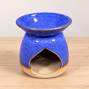 May include: A ceramic wax warmer featuring a vibrant blue speckled glaze on the upper section and a wide, bowl-shaped top. The base is rounded with a large, arched opening in the front, revealing a natural, unglazed tan clay interior surface.