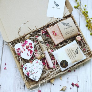 Può includere: Una scatola regalo piena di prodotti da bagno, tra cui un sapone al sale marino di rosa, bombe da bagno e sali da bagno. La scatola è piena di carta triturata e decorata con petali di rosa secchi.