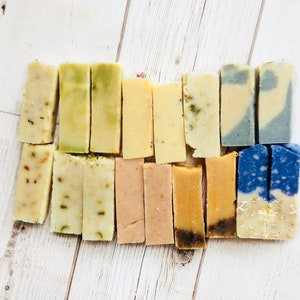 May include: A collection of 16 handmade soap bars in various colours and patterns, including green, yellow, blue, and pink. The bars are arranged in two rows on a white wooden surface.