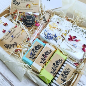 May include: A gift box filled with handmade bath and body products. The box contains a variety of soaps, bath melts, and a facial steam. The products are all natural and feature a variety of floral scents. The box is decorated with a hand-made label and a sprig of rosemary.