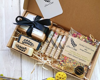 Puede incluir: Una caja de regalo llena de productos naturales para el baño y el cuerpo. La caja contiene dos barras de jabón, cinco pequeños frascos de vidrio llenos de sales de baño y una bolsa de hierbas secas para un baño de vapor facial. La caja está decorada con una cinta negra y una pequeña etiqueta con el texto "HeniGoap".