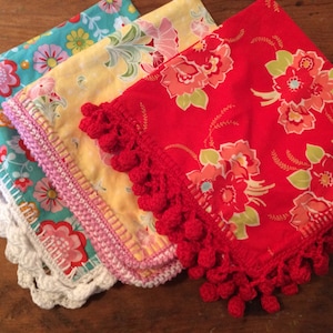 May include: Three fabric napkins with crocheted edges. The napkins are in different floral patterns: turquoise with white flowers, yellow with pink flowers, and red with pink and white flowers. The crocheted edges have pom-poms.