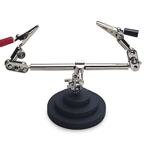 May include: A silver soldering station with two adjustable alligator clips, a black base with three tiers, and red and black insulated wire ends.