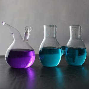 May include: Three glass beakers with a round bottom. The beaker on the left is purple, the two beakers on the right are blue.