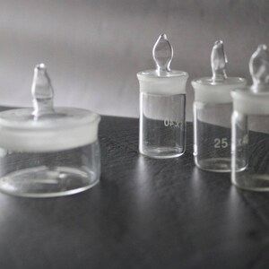 May include: Four clear glass bottles with stoppers. The bottles are of different sizes and have markings on the side. The bottles are on a dark grey surface.