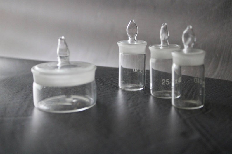 May include: Four clear glass bottles with stoppers. The bottles are of different sizes and have markings on the side. The bottles are on a dark grey surface.