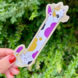 May include: A quirky cat sticker with a cream border. The cat features a white body with purple, yellow, and black spots. The cat is in a playful pose with its paws raised. The background is a blurred green.
