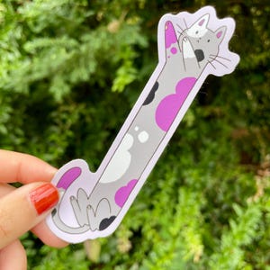 May include: A whimsical sticker featuring a long, stylized cat in a playful pose. The cat has a gray body with white, black, and pink heart-shaped markings. The sticker is held against a backdrop of green foliage.
