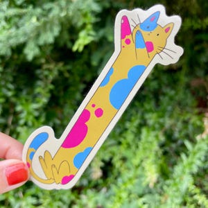 May include: A whimsical sticker featuring a yellow cat with a long body, blue and pink spots, and a blue hat. The cat is in a playful pose, with its paws raised. The sticker has a white border and is set against a blurred green background.