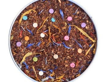May include: A round tin filled with loose leaf tea. The tea is a blend of brown, orange, and blue pieces. There are small, colourful sweets scattered throughout the tea.