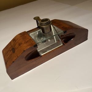 May include: A dark brown wooden inkwell stand featuring a central glass reservoir and a metallic brass-toned cap. The base has a smooth, polished finish with two carved side sections and a rectangular glass insert surrounding the central ink pot.