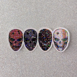 May include: Four skull stickers with glitter accents. Each skull has a unique color scheme: red, black, dark purple, and a mix of white and orange. The stickers are on a silver glitter background.