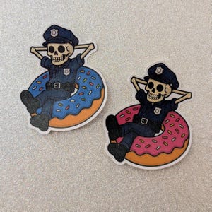 May include: Two stickers featuring a skeleton police officer relaxing on a donut. The officer wears a blue uniform and hat, with arms behind the head. One donut is blue with sprinkles, the other is pink.