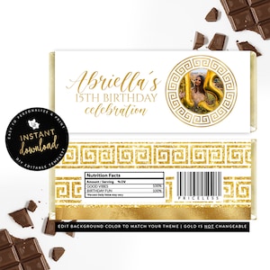 May include: Gold and white printable chocolate bar wrapper with a Greek key pattern and the text "Abriella's 15th Birthday celebration". The wrapper also features a photo of a person holding a gold balloon number 15.