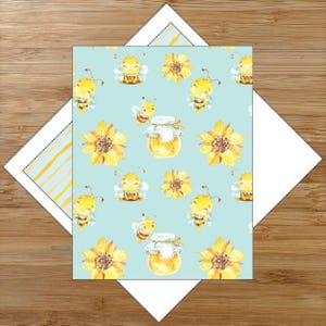 May include: A greeting card with a light blue background featuring a pattern of smiling cartoon bees, sunflowers, and honey jars. The card is surrounded by a white envelope and a striped blue and yellow insert.