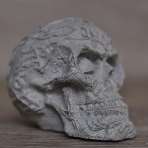 May include: A detailed, gray, concrete skull sculpture with intricate carvings and a textured surface.